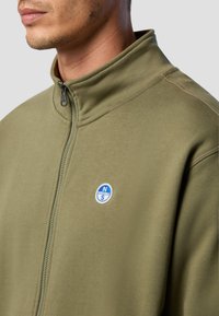 Giacca in pile verde oliva con zip e colletto alto, caratterizzata da un logo circolare blu e bianco sul petto e dettagli delle cuciture ben visibili.