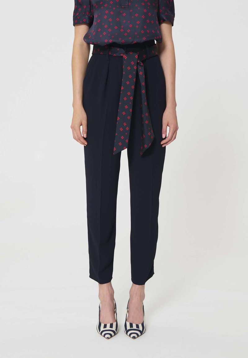 Femme portant un pantalon taille haute bleu marine avec une ceinture nouée à motifs, associée à un haut bleu marine assorti et des talons hauts rayés noir et blanc.