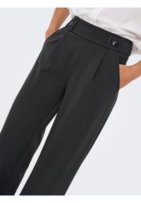 Pantalon noir taille haute avec plis devant et taille boutonnée, porté avec les mains dans les poches et une chemise blanche rentrée visible au-dessus.