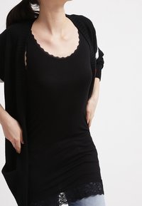 Svart linne med en scalloped spetskant. Har en rundad halsringning och en figurnära silhuett, kompletterad av en lager-på-lager, öppnad svart kofta.