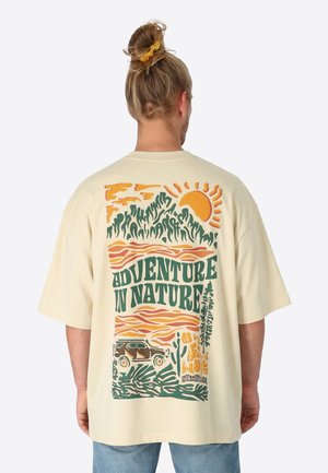 Blonde Person mit zusammengebundenem Haar, der ein cremefarbenes Shirt trägt und eine bunte Grafik mit der Aufschrift "Abenteuer in der Natur" zeigt, auf der Sonne, Berge, Bäume und ein Fahrzeug zu sehen sind.