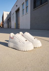 Baskets à plateforme blanches avec semelles épaisses et accents rose pâle placées sur du béton ensoleillé près d'une rangée de bâtiments industriels.