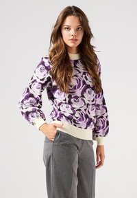 Maglione a fantasia floreale viola con collo e polsini a coste crema, abbinato a pantaloni grigi, che mostra una vestibilità rilassata e una texture morbida.