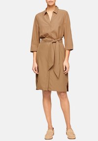 Vestito marrone con bottoni, vita con lacci, maniche a tre quarti, spacchi laterali e una consistenza liscia. Abbinato a scarpe beige chiare senza lacci.