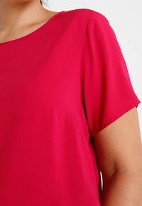 Top a maniche corte e scollo tondo in tessuto di seta fucsia. Presenta una consistenza morbida e liscia con maniche leggermente a sbuffo. Dettagli delle cuciture visibili.