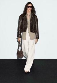 Bruine leren cropped jack, beige shirt en losse witte broek. Gecombineerd met een bruine handtas en donkere oversized zonnebril.