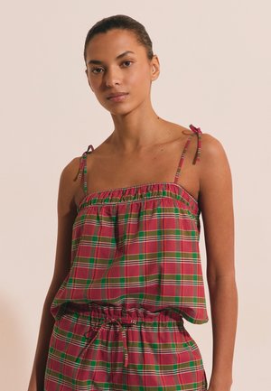 Młoda kobieta ubrana w bezrękawnik w czerwono-zieloną kratę z cienkimi ramiączkami, stojąca na jednolitym beżowym tle.