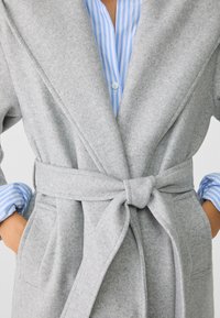 Mujer vistiendo un abrigo de lana gris claro con cinturón sobre una camisa de botones azul y blanca a rayas, con las manos en los bolsillos del abrigo.