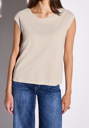 Kvinde iført beige top med korte ærmer og blå jeans, stående med armene afslappet ned langs siderne mod en ensfarvet baggrund.