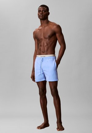 Kaal bovenlichaam mannelijke model draagt lichtblauwe zwembroek met witte Calvin Klein tailleband, staat blootsvoets tegen een effen grijze achtergrond.