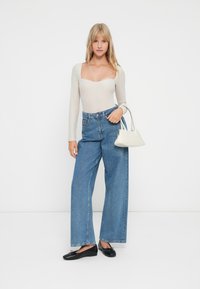 Helles beiges Langarmoberteil mit rechteckigem Ausschnitt, kombiniert mit lockeren blauen Jeans. Schwarze Loafer und eine kleine weiße Handtasche runden das Outfit ab.