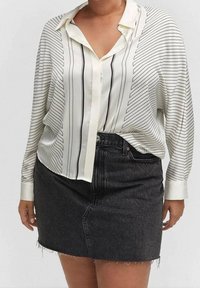 Weiße Bluse mit schwarzen vertikalen Streifen, ausgestattet mit einem Kragen und langen Ärmeln. Kombiniert mit einem schwarzen Jeans-Minisrock mit fransigem Saum.