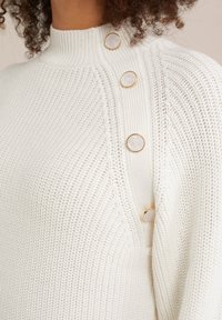 Baltas mezginių džemperis su aukštu kaklu, tekstūruotu audiniu ir keturiais aukso akcentuotais mygtukais ant peties, suteikiančiais papildomą detalę.