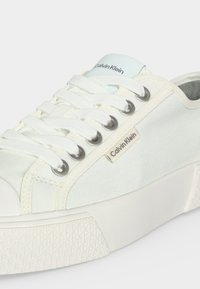 Baskets basses blanches Calvin Klein avec lacets blancs, œillets métalliques, semelle texturée et étiquettes de la marque sur le côté et la languette.
