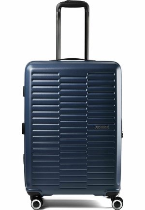 Hartschalenkoffer in Marineblau mit Teleskopgriff und vier Rollen, vorne mit horizontalem Rillenmuster, American Tourister-Logo auf der Vorderseite.