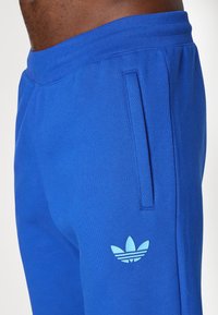 Zilas sportiskas bikses, kas izgatavotas no mīkstas auduma, ar šauru piegriezumu, sānu kabatu un gaiši zilu Adidas logotipu kreisajā augšstilbā.