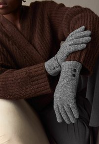 Graue Wollhandschuhe mit glatter Textur haben einen Bund mit zwei schwarzen Knöpfen und werden mit einer gemütlichen braunen Strickjacke kombiniert.