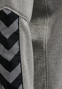 Giacca sportiva grigia caratterizzata da un tessuto testurizzato con un pannello di materiale nero decorato con strisce a forma di chevron bianche sulla manica.