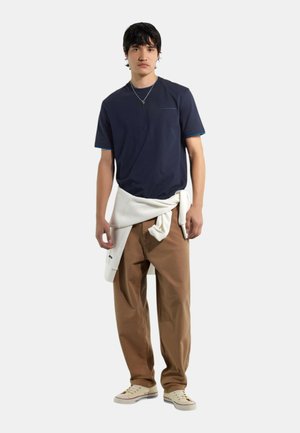 Giovane uomo che indossa una t-shirt blu navy, pantaloni beige, scarpe da ginnastica bianche e una felpa bianca legata in vita, in piedi davanti a uno sfondo semplice.