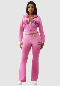 Veste à zip rose avec imprimé graphique et pantalon évasé assorti. Tissu doux, silhouette ajustée et accents côtelés. Des baskets blanches complètent le look.