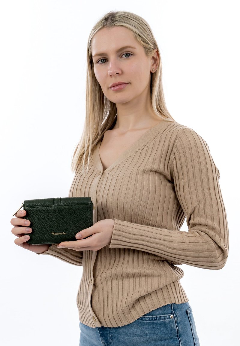 Femme aux longs cheveux blonds portant un pull beige côtelé et un jean bleu, tenant un portefeuille en cuir vert foncé avec fermeture éclair.