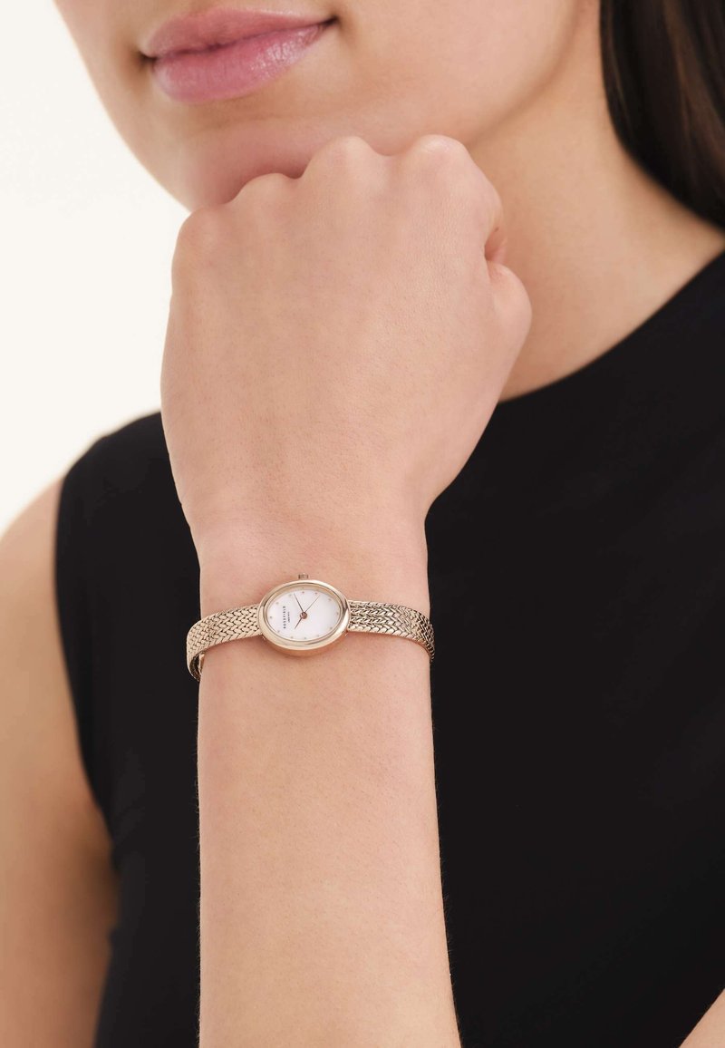 Femme portant une montre-bracelet à maille dorée avec un cadran ovale blanc à son poignet gauche, sur un fond de haut noir sans manches.