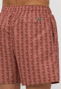 Kupaći šorts s uzorkom u rustikalno crvenoj boji, s geometrijskim dizajnom i elastičnom strukom. Značajan Lacoste logo u zelenoj boji na stražnjem džepu.