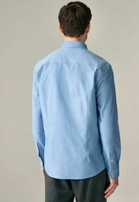 Camisa azul clara de botones en una tela suave con mangas largas, pliegues en la espalda y un dobladillo redondeado. Presenta un cuello estándar y puños con botones.