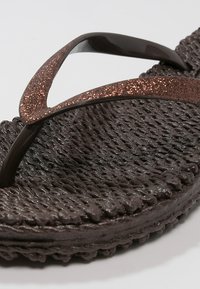 Gros plan d’une tong marron foncé texturée avec des brides marron pailletées sur un fond clair.