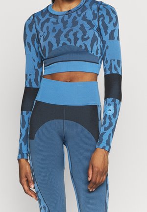 Kobieta ubrana w niebiesko-czarny, wzorzysty długi crop top z długim rękawem oraz dopasowane legginsy z wysokim stanem, stojąca na jednolitym tle.