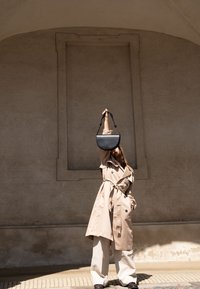 Personne portant un trench beige et un pantalon blanc large lève un sac à main noir au-dessus de la tête sur un mur texturé en arrière-plan.