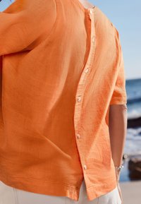 Lyhythihainen oranssi pellavapaita, jossa on napitus selän takana ja rento istuvuus, ranta-taustaa vasten.