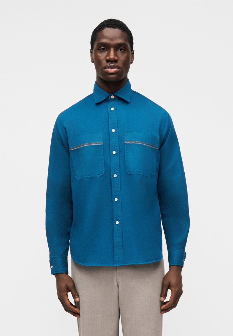 Τυρκουάζ πουκάμισο με κουμπιά, δύο τσέπες στο στήθος, ίσιο κάτω μέρος και μακριά μανίκια. Υφή υφάσματος με λεπτομέρειες αντίθετης ραφής.