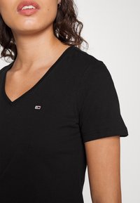 Czarna bawełniana koszulka z dekoltem w serek, krótkimi rękawami i małym haftowanym logo na górnej lewej stronie. Gładka tekstura.