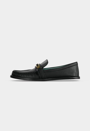 Mocassin noir en cuir avec un détail de boucle dorée sur la lanière, semelle plate et coutures style mocassin sur l’avant.