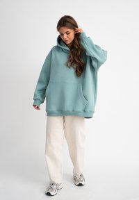 Felpa oversized color teal realizzata in morbido tessuto, con una grande tasca frontale e polsini a coste, abbinata a pantaloni color crema e sneakers.