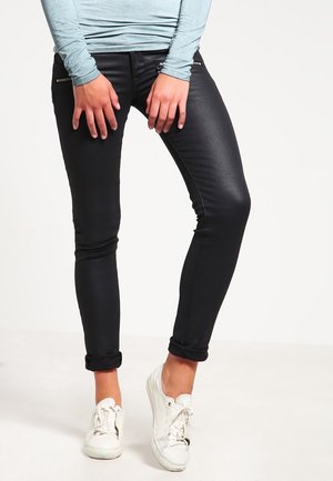 Persona vistiendo pantalones negros de corte ajustado con puños enrollados, una camiseta de manga larga azul claro y zapatillas blancas, de pie frente a un fondo liso.