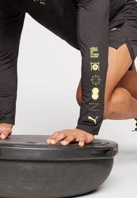 Černé sportovní tričko s dlouhým rukávem a žlutou grafikou na rukávu, obsahující symboly a text. Ruce spočívají na černé kulaté cvičební plošině.