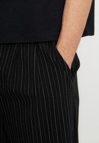 Δεξί χέρι στην τσέπη μαύρου παντελονιού με ρίγες, φορώντας μαύρο πουκάμισο, εμφανίζεται από τη μέση μέχρι το μέσο του μηρού σε απλό φόντο.