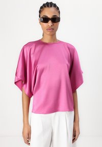 Fuksia satintopp med brede ermer og rund hals, kombinert med hvite bukser. Modell med svarte rektangulære solbriller.