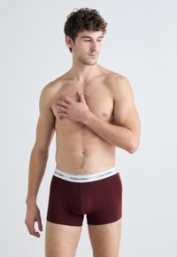 Burgundi Calvin Klein boxer alsónemű, fehér márkás derékszíjjal. Simasága, testhez álló fazonja és a comb közepéig érő hossza jellemzi.