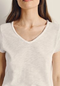 Sieviete valkā baltu īss piedurkņu V-veida kakla kreklu ar neapstrādātām malām, redzams kakls, atslēgas kauls un daļa no garajiem brūnajiem matiem.