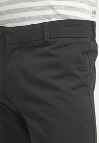 Svarta byxor med flat front, sidfickor och bälteshällor. Tyget ser slätt ut med en subtil lyster. Presenteras med en grårandig skjorta.