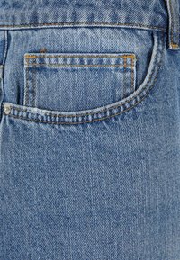 Jean en denim avec une poche arrière, de couleur bleu clair, avec des coutures distinctes en orange et une finition lisse et texturée.