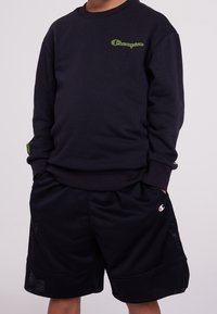 Black sweatshirt with green "Champion" logo, paired with black mesh shorts featuring logo accents. Casual sportswear design and fit.