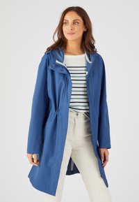 Capa de agua azul con cintura ajustable, capucha y cierre delantero a presión. Llevada sobre una camisa a rayas blanca y azul, combinada con pantalones claros.