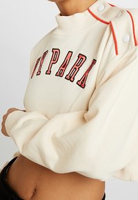 Mikina s kulatým výstřihem v krémové látce s červeným a černým nápisem "IVY PARK", žebrovaným límcem a detaily na rameni zdobenými knoflíky.