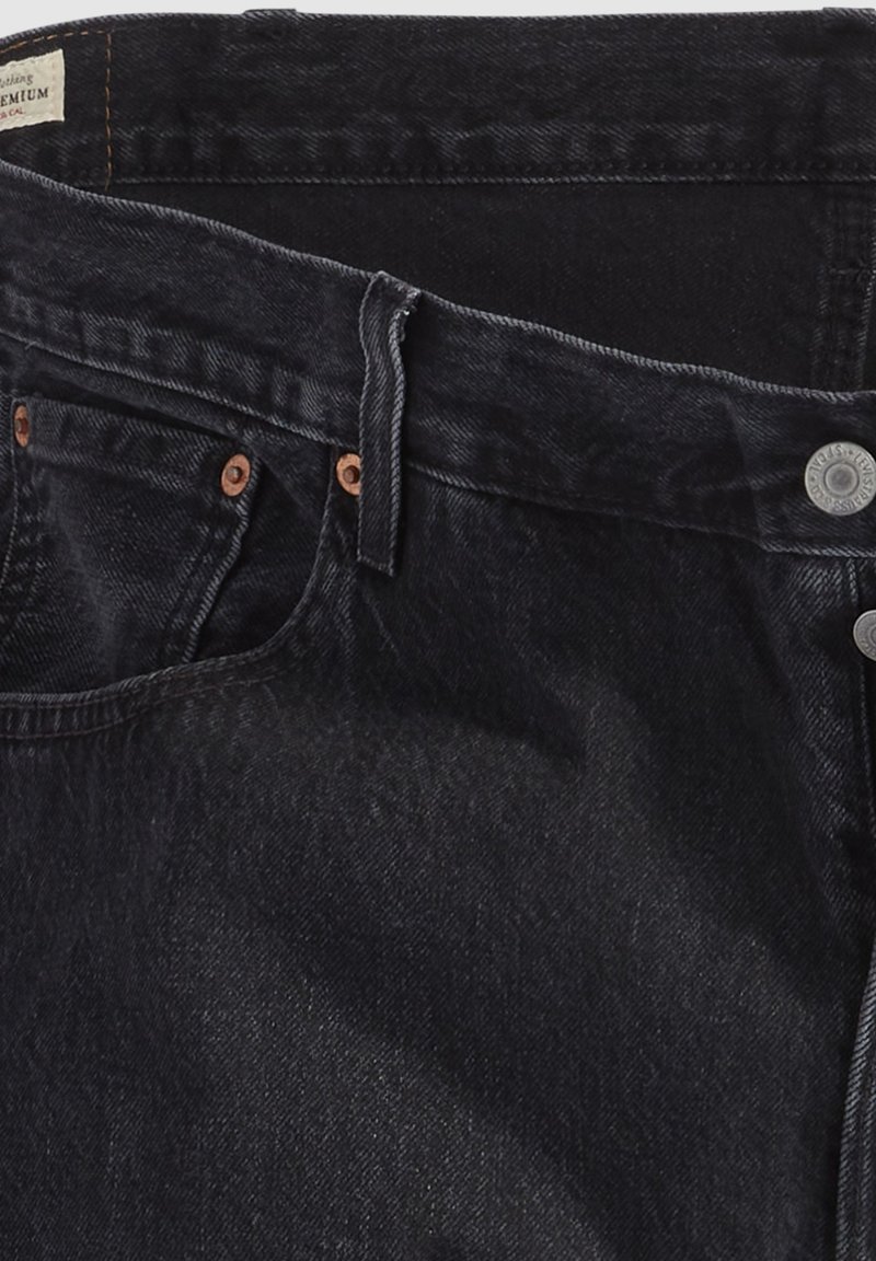 Jean en denim noir avec une texture lisse, ornés de rivets en cuivre et d'une fermeture à bouton. La taille est équipée d'une étiquette et de passants de ceinture.