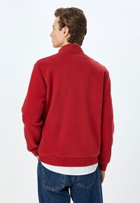 Felpa rossa con colletto alto, realizzata in tessuto morbido. Presenta polsini e orlo a costine, indossata sopra una camicia bianca e jeans blu larghi.