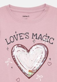 Camiseta de algodão rosa com um grande design de coração com um acabamento brilhante, cercado pelo texto "A MAGIA DO AMOR" e pequenos elementos decorativos.